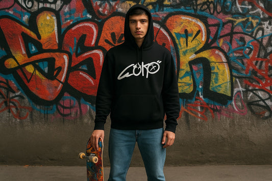 Young man in black hoodie with bold white Geko graffiti logo, holding a skateboard, standing in front of a colourful graffiti wall in an urban streetwear setting.